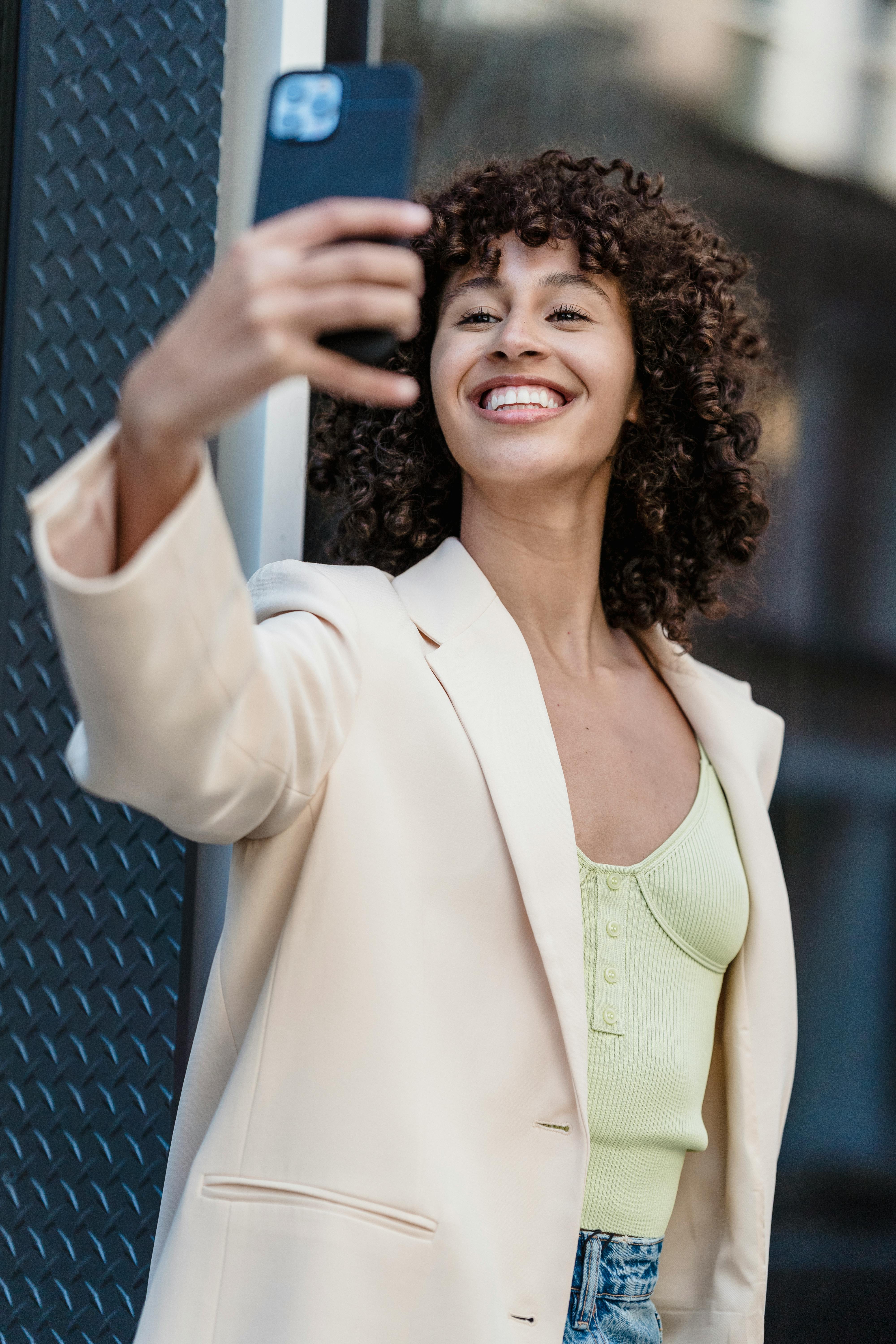 Cheerful young ethnic female with curly hair in jacket taking self portrait on cellphone on street