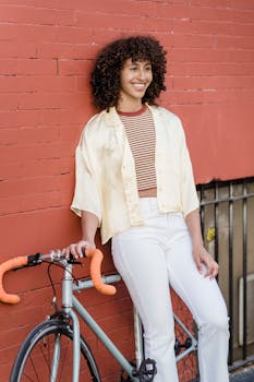 Cheerful woman standing with bicycle against a red brick wall, embracing urban style.
