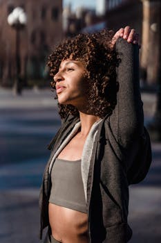Contemplative young ethnic female athlete with curly hair warming up while looking away in evening town
