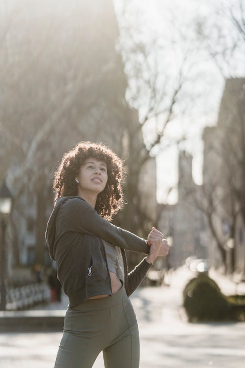 Concentrated ethnic female athlete with Afro curls wearing sportswear stretching arms and listening to music in wireless earbuds during fitness training in park