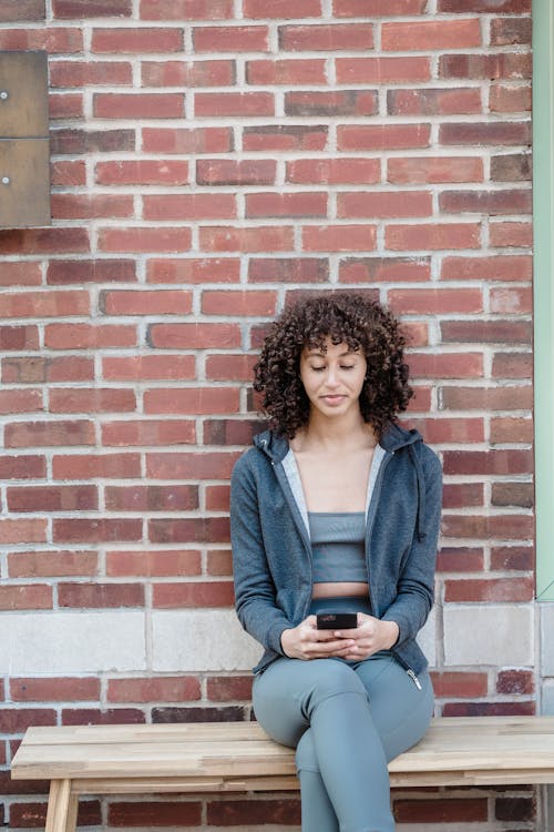Ethnic curly haired sportswoman browsing smartphone on bench