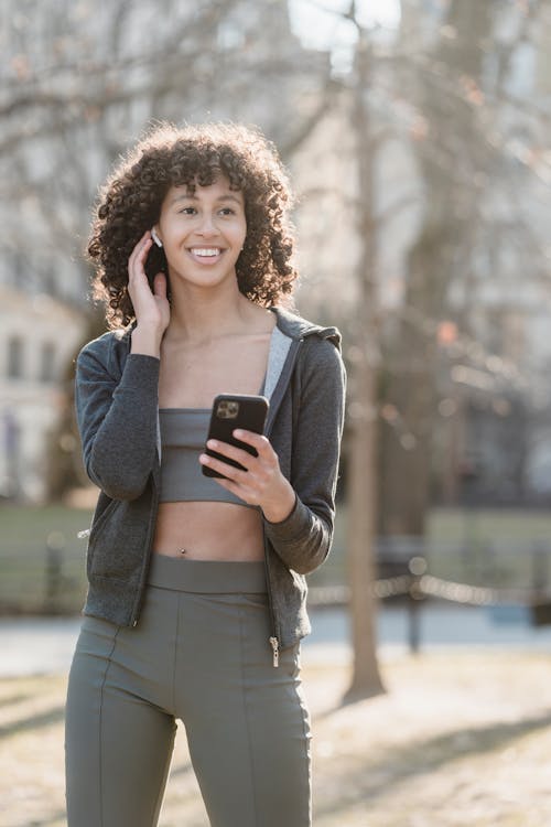 Free Positive young ethnic sportswoman in activewear smiling happily while communicating by wireless earbuds connected with cellphone Stock Photo