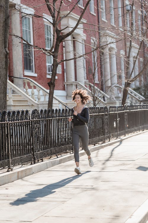 Free Full body of positive ethnic female with flying curly hair running on walkway along city buildings during fitness training Stock Photo