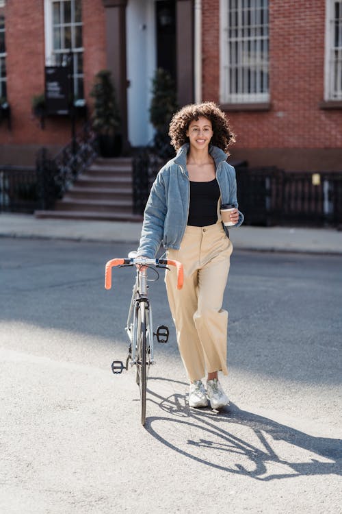 Full body of smiling ethnic female with cup of takeaway beverage crossing road with bicycle