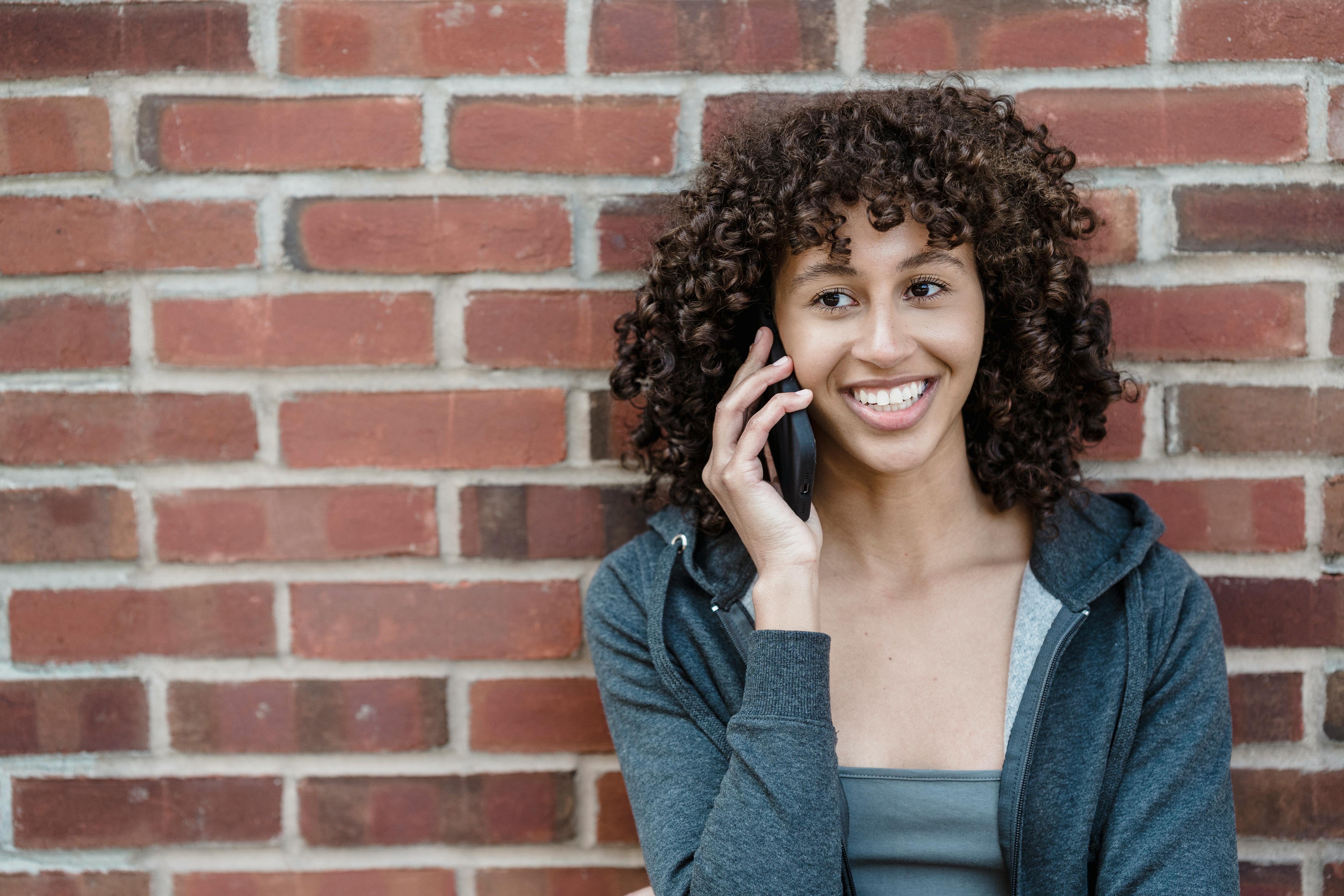 smiling ethnic woman speaking on smartphone near brick wall