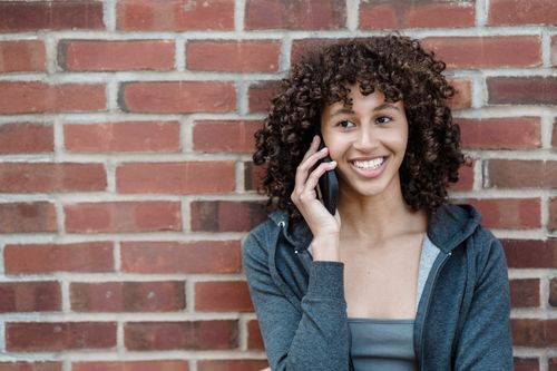 Free Positive young ethnic female with curly dark hair smiling happily while having phone conversation against brick wall Stock Photo