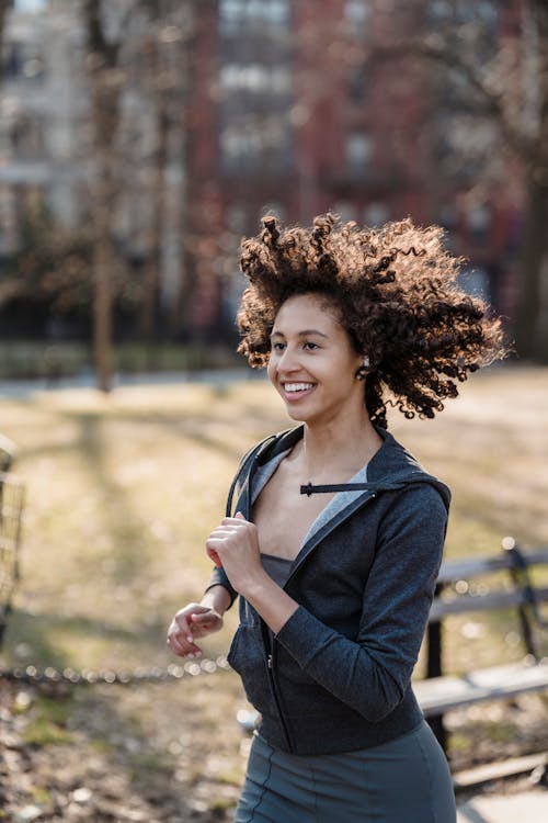 Cheerful ethnic female runner with flying hair listening playlist in wireless earbuds while jogging in city park