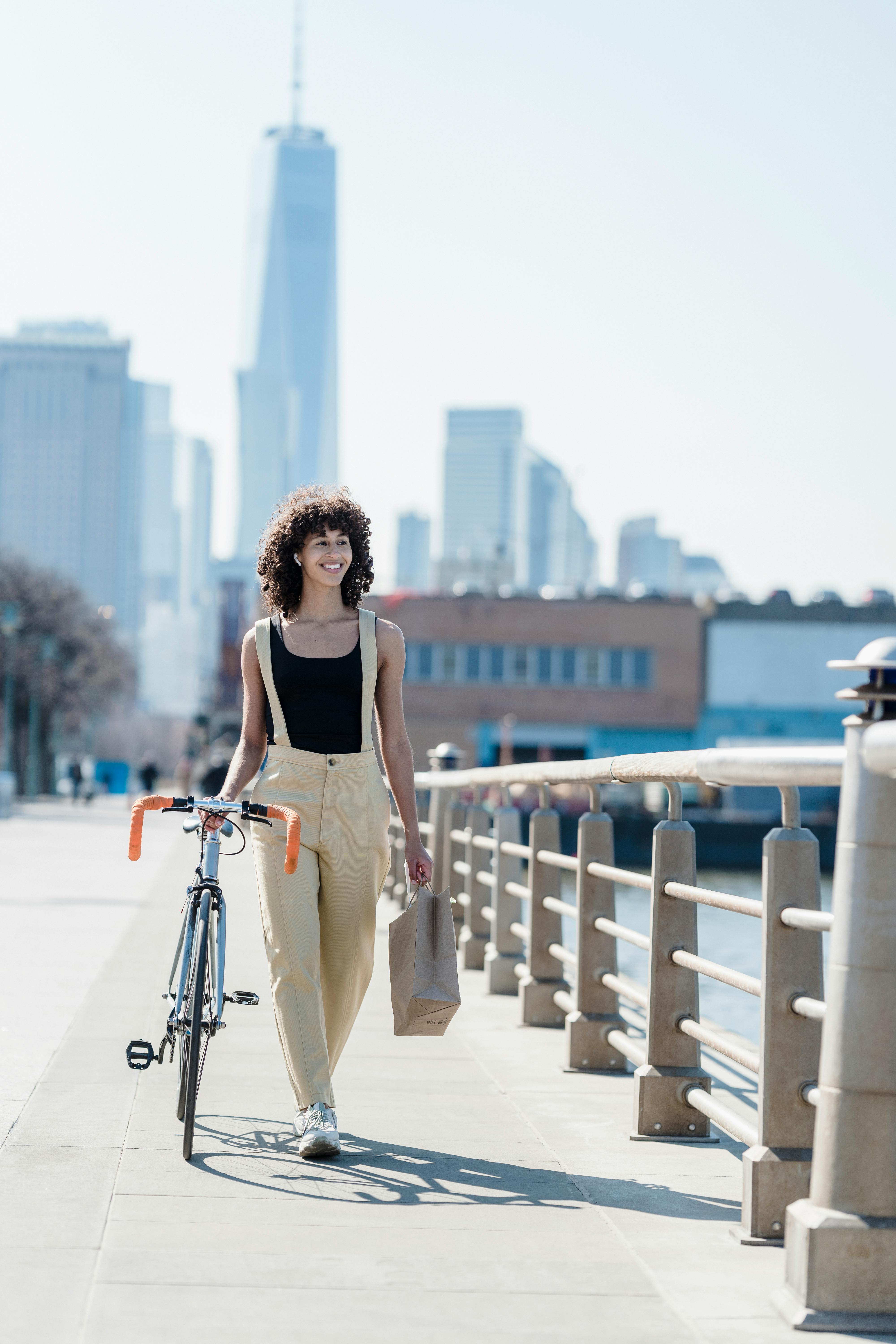 Free Full body of positive ethnic female listening to music in wireless earbuds while strolling with bicycle and paper shopping bag on waterfront Stock Photo