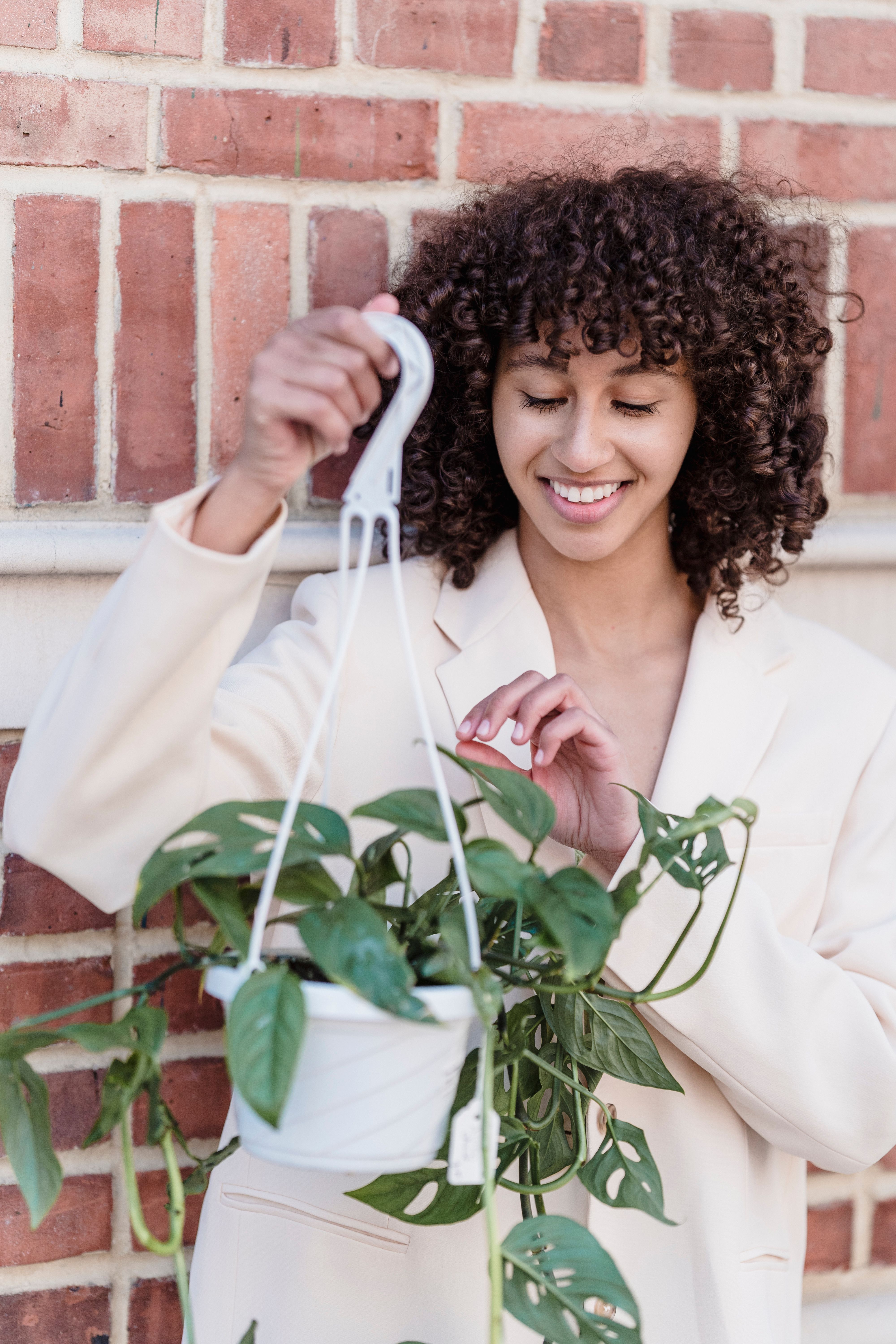 Free Positive ethnic female with curly hair standing with houseplant in hanging planter near brick wall Stock Photo