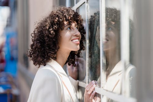 Side view of positive ethnic female with curly hair standing near window and looking up with interest
