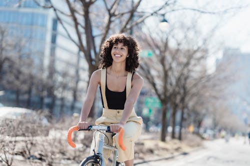 Cheerful active ethnic female cyclist looking away while riding bicycle on walkway on sunny street with residential buildings and trees