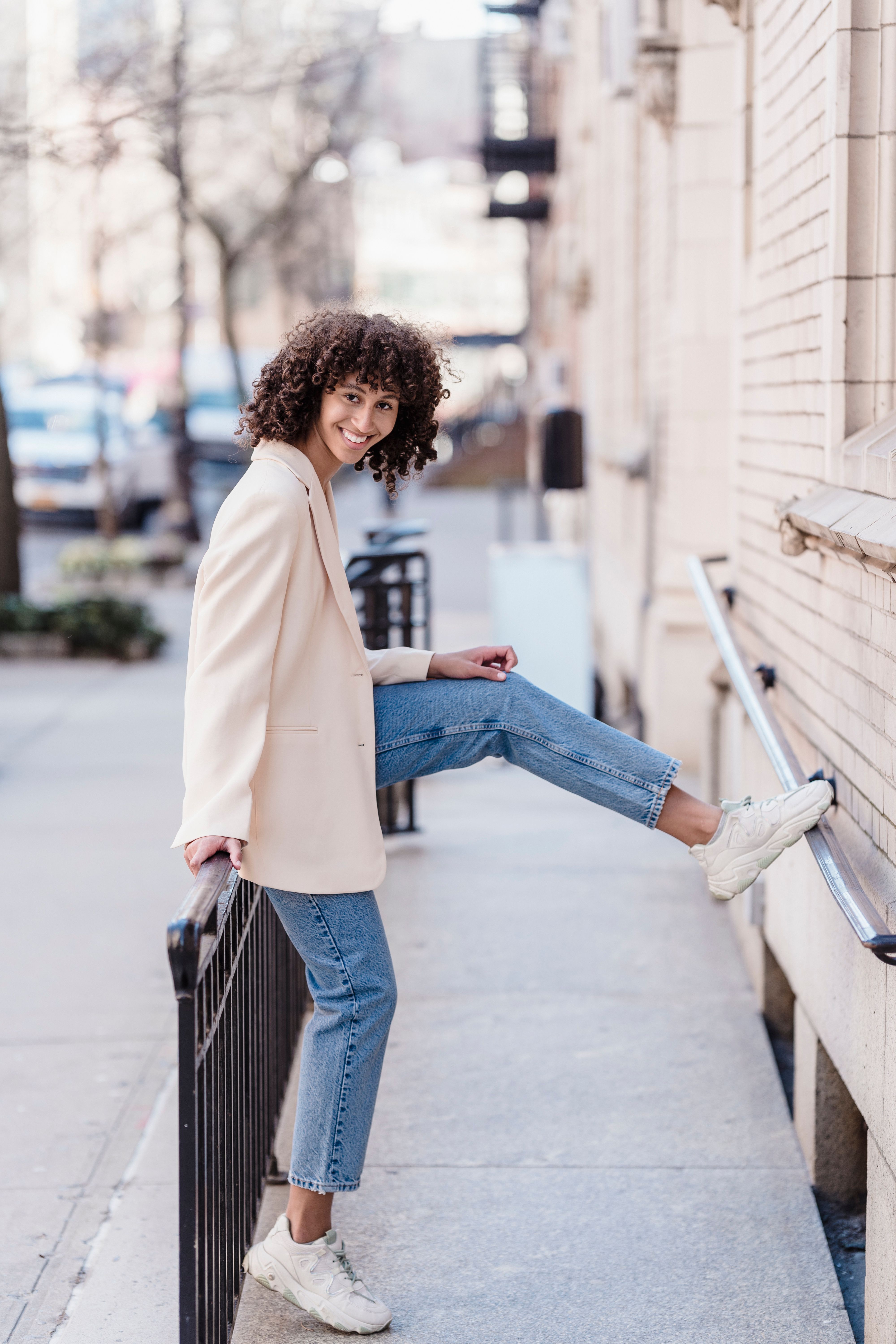 Free Full body of cheerful ethnic female in trendy outfit looking at camera while standing on walkway with raised leg near building Stock Photo