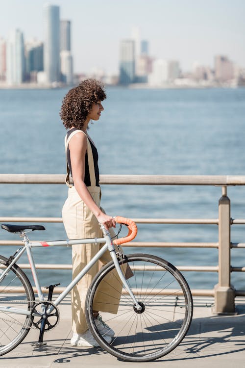 Full body side view of ethnic female with bicycle standing on waterfront with metal fence near rippling sea against modern buildings in city