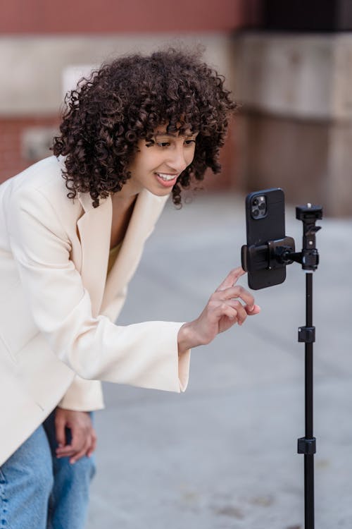Content ethnic female blogger in stylish wear touching screen of smartphone while recording live video on walkway near building in city