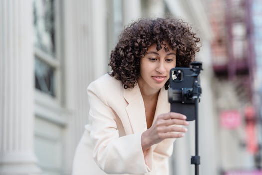 Woman in a cream blazer adjusting a smartphone on a stand, photographed outdoors.