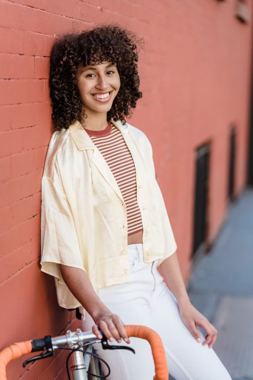 Free A cheerful woman with curly hair leans against a brick wall, holding a bicycle, outdoors. Stock Photo