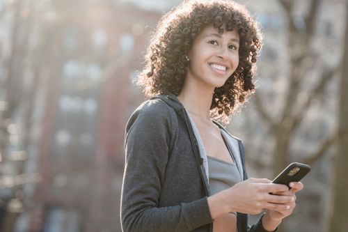Free Woman with curly hair using smartphone outdoors, smiling Stock Photo