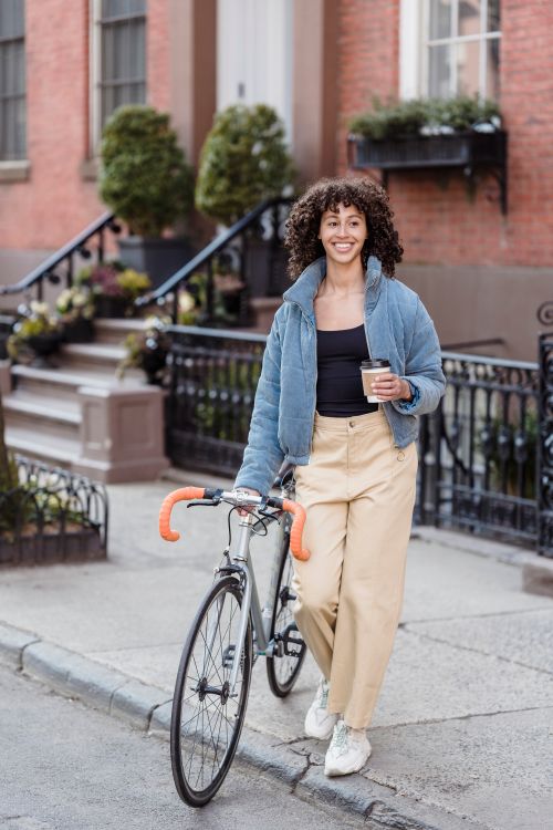 Free Smiling young woman walking on street with bicycle and holding a cup of coffee. Stock Photo