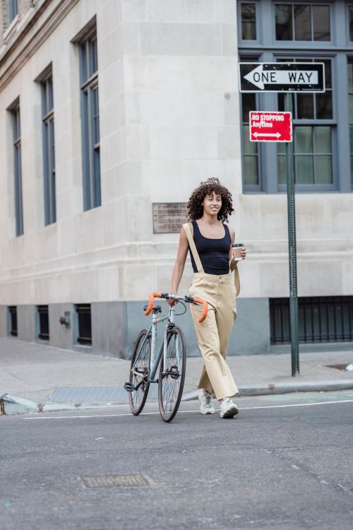 Free Young woman walking a bicycle on an urban street, holding a coffee cup. Stock Photo