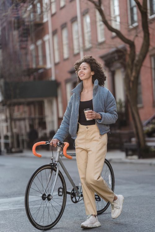 Free A young woman in casual attire holds coffee and walks with a bicycle on a city street. Stock Photo