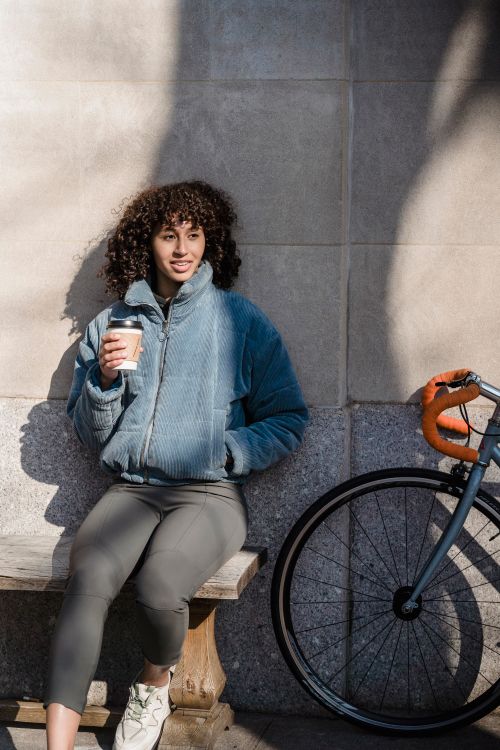 Free A young woman sits on a bench holding a coffee in a sunny urban street scene. Stock Photo