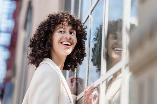 Free A cheerful woman with curly hair smiles by a city building, reflecting in glass. Stock Photo