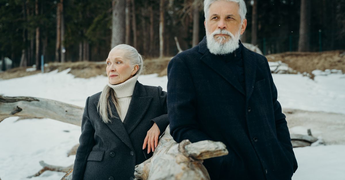Senior couple wearing winter clothes leaning on tree log in snowy forest setting.