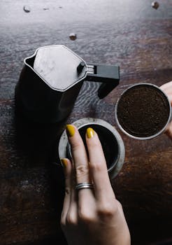 A person assembling a Moka pot with freshly ground coffee, showing hands and coffeepot on wooden surface.