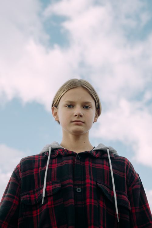 Free A teenager in a plaid hoodie poses outdoors against a blue sky, looking confidently at the camera. Stock Photo