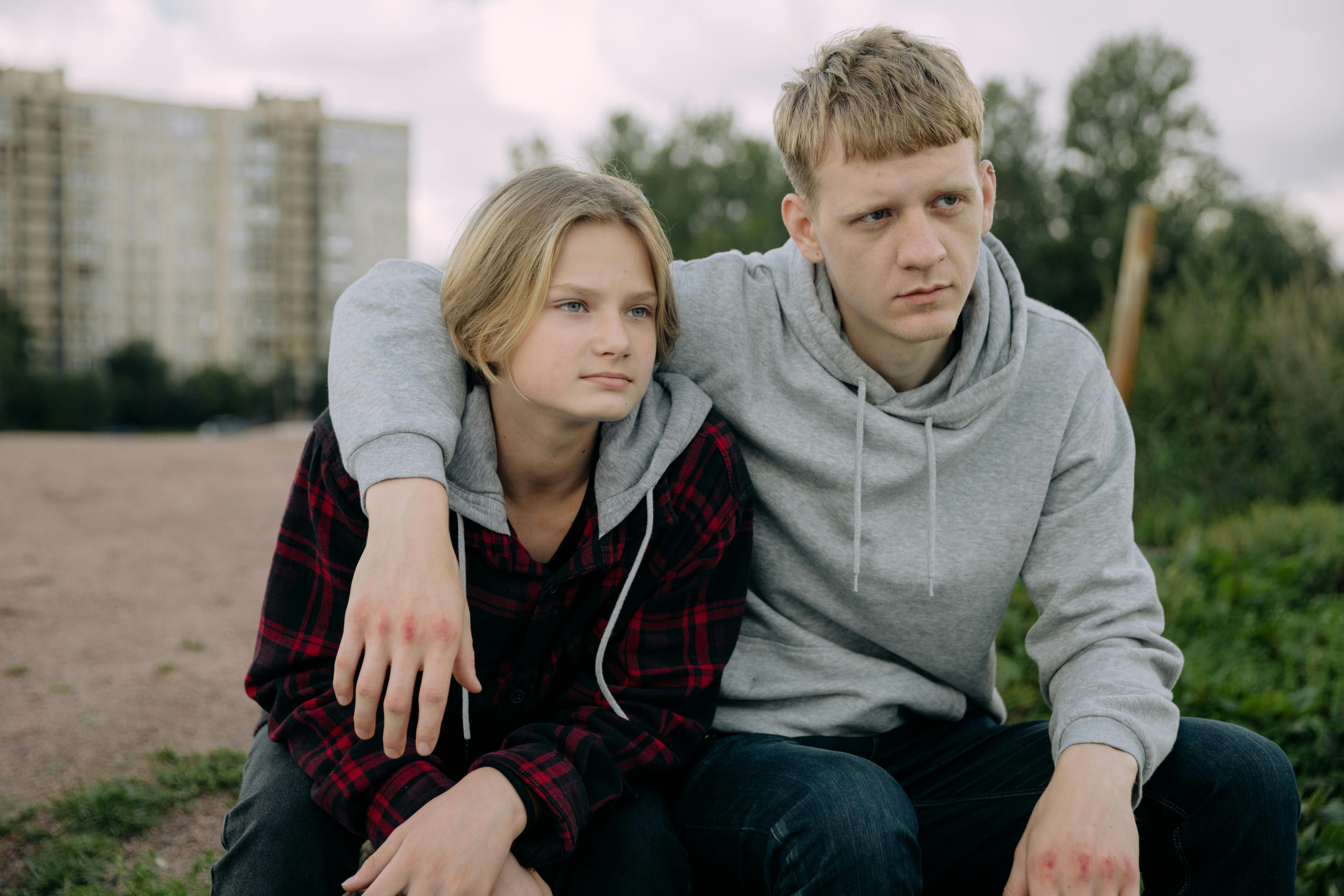 Two siblings in casual hoodies sit together outdoors, conveying a relaxed and thoughtful moment.
