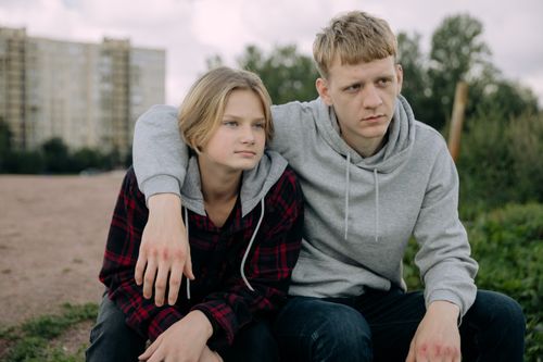 Free Two siblings in casual hoodies sit together outdoors, conveying a relaxed and thoughtful moment. Stock Photo