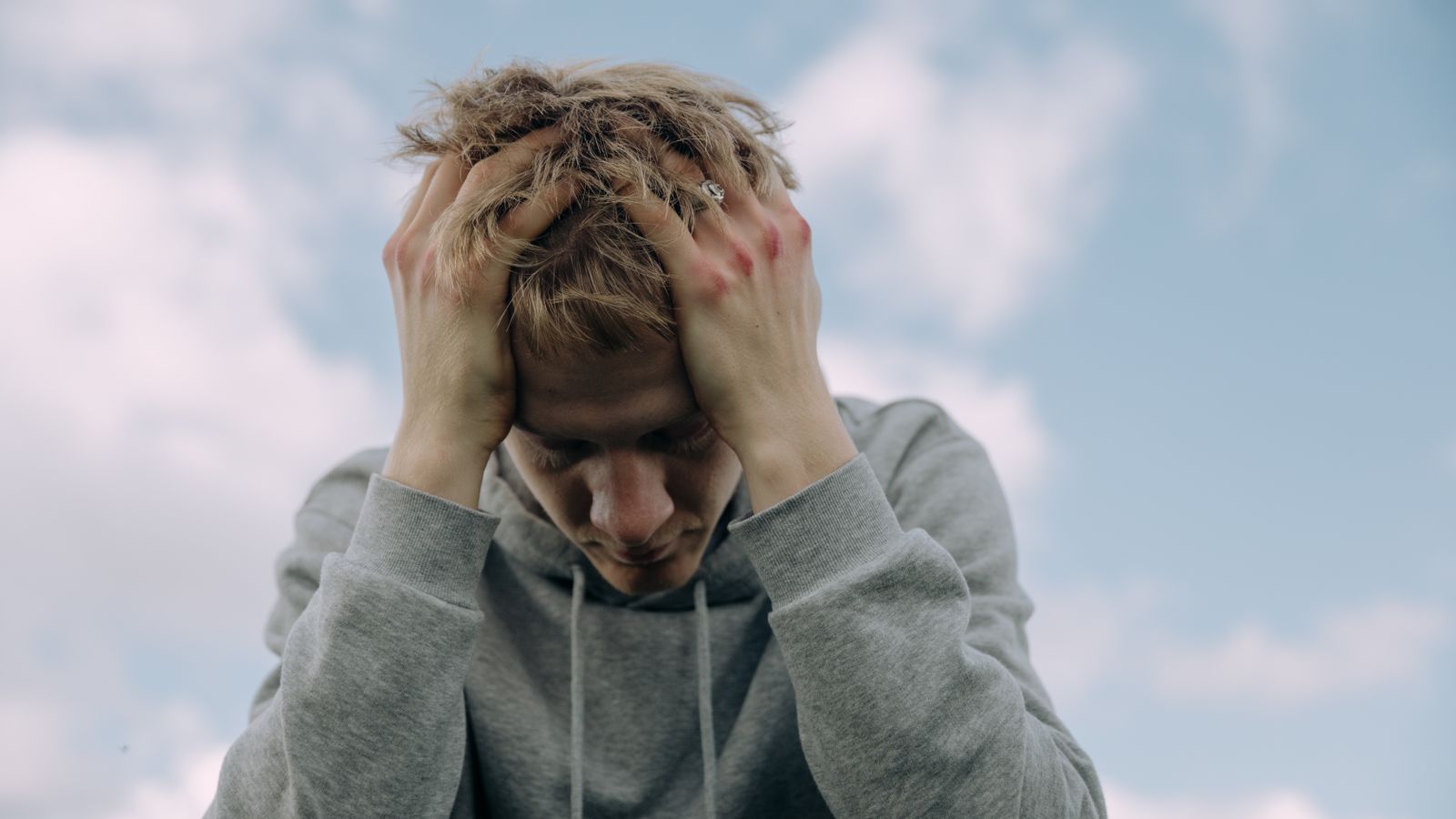 Young man in gray hoodie holds head in frustration, set against cloudy sky.