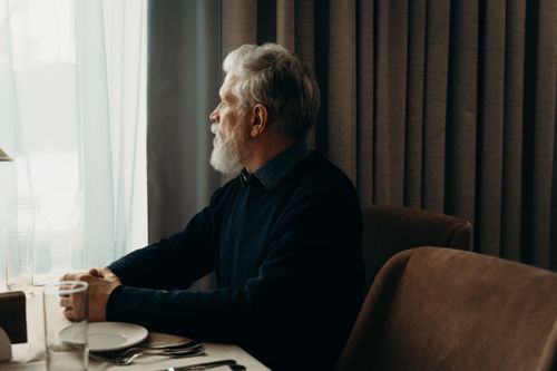 Free An elderly man with a beard looking outside while sitting by a table indoors. Stock Photo