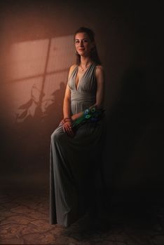 A serene portrait of a woman in a long gray dress holding peacock feathers in a dimly lit room.