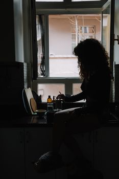 Silhouette of a woman sitting by a window in a kitchen, creating a dramatic mood.
