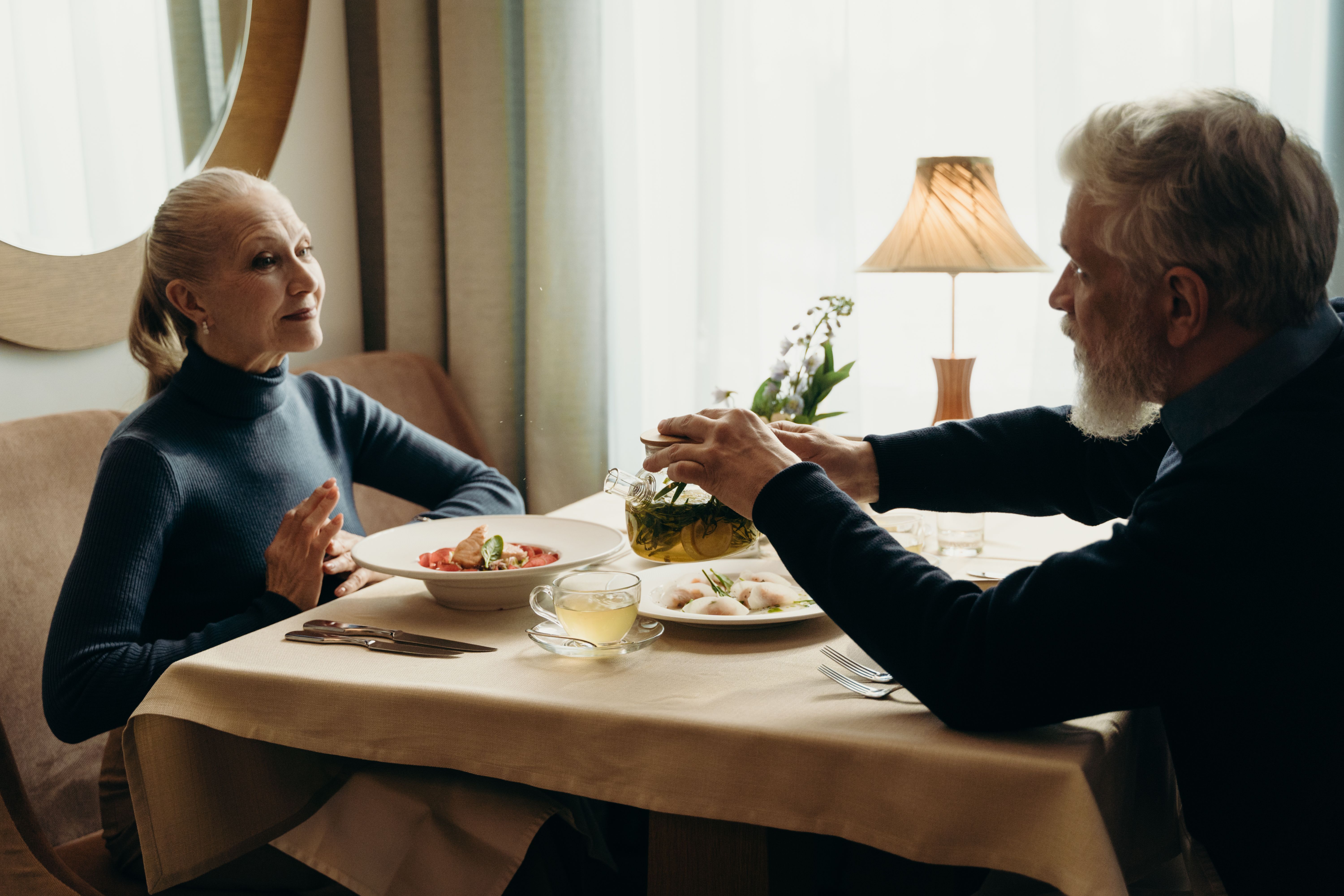 A senior couple dining and pouring tea in a cozy indoor setting, emphasizing warmth and companionship.