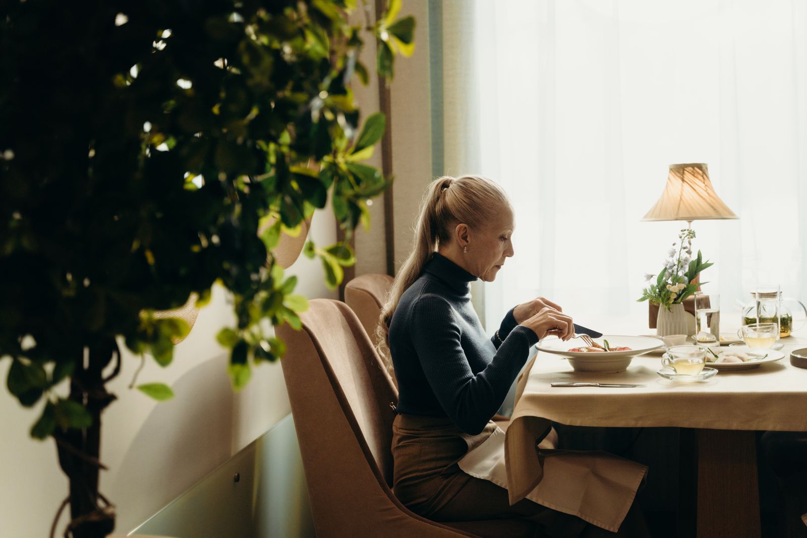 Woman with blonde ponytail in black turtleneck eating at a cream-draped table beside a sunlit window
