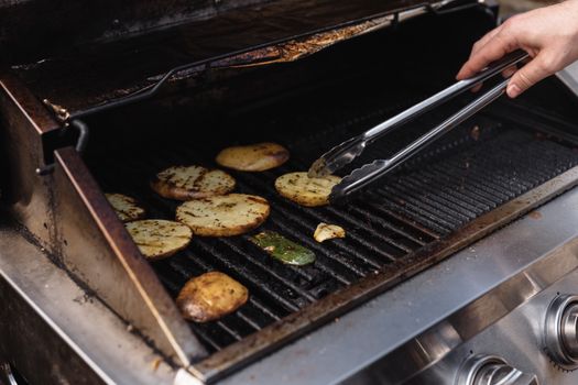 Unrecognizable man with tongs turning over tasty cut potatoes on metal grate while cooking on modern hot barbecue grill machine