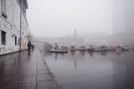 Many boats moored on calm water surface near paved waterfront with people walking near shabby building in town on foggy weather