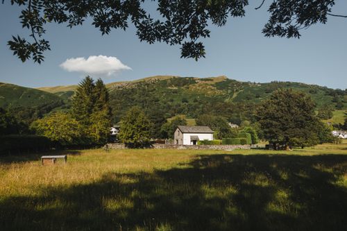 Free White residential cottage located on grassy ground near tall green trees and mountain ridge covered with grass in rural area Stock Photo