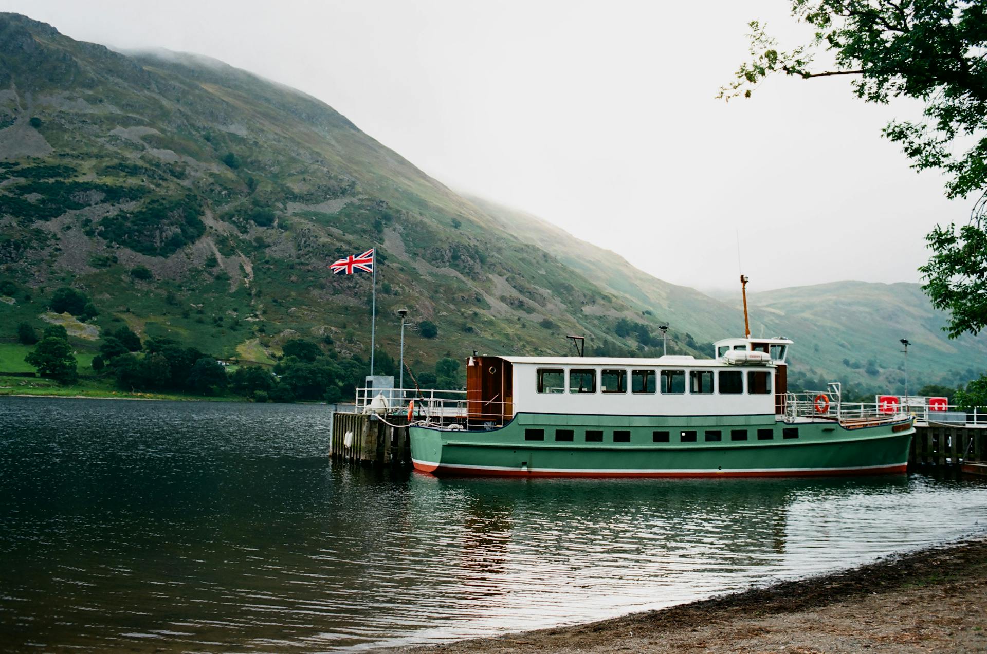 Passenger ferry with flag of England moored on rippling water near coast against green hills covered with grass in nature