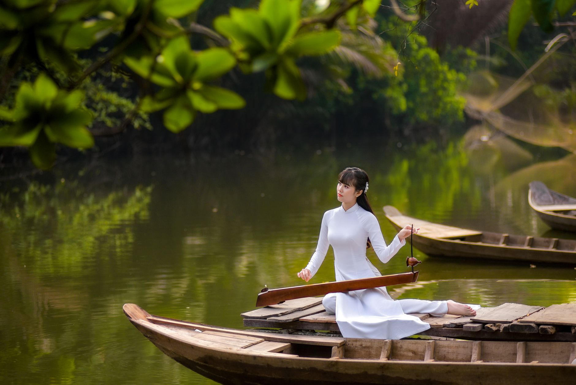 Woman in white dress playing Dan Bau on a boat amid lush greenery.