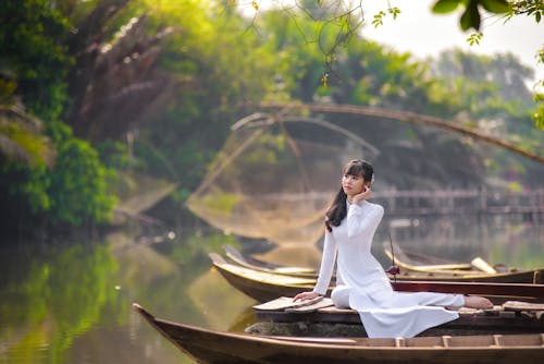 Selective Focus Photo of a Woman in a White Dress Sitting Near a Boat