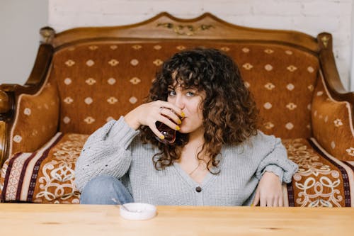 Woman in a Gray Sweater Leaning on a Brown Sofa While Drinking Turkish Tea