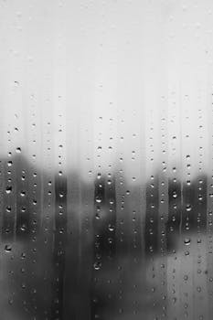 Close-up of raindrops on a glass window in grayscale, creating a moody atmospheric effect.