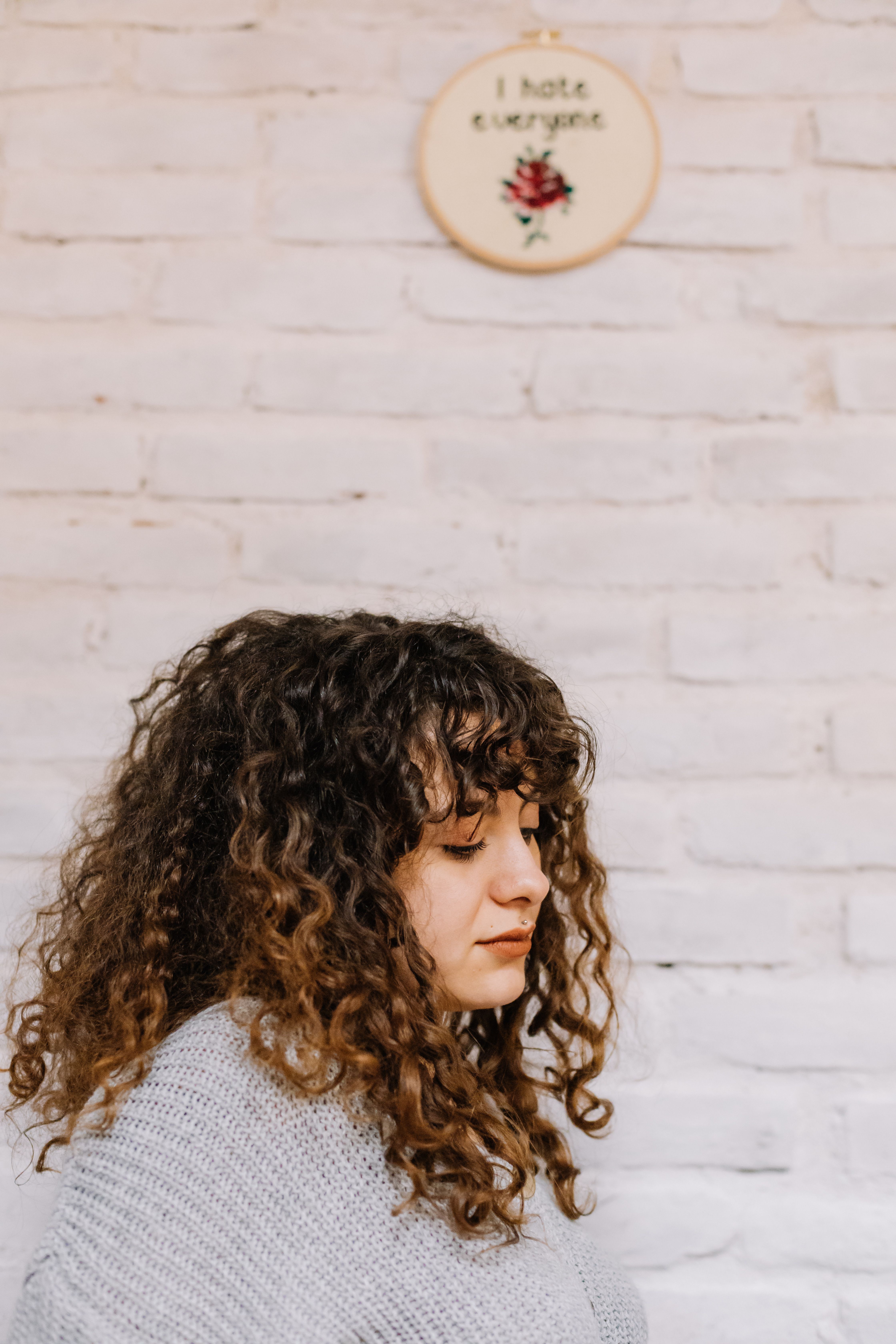 Profile of a young woman with curly hair against a white brick wall with a humorous embroidery art piece.