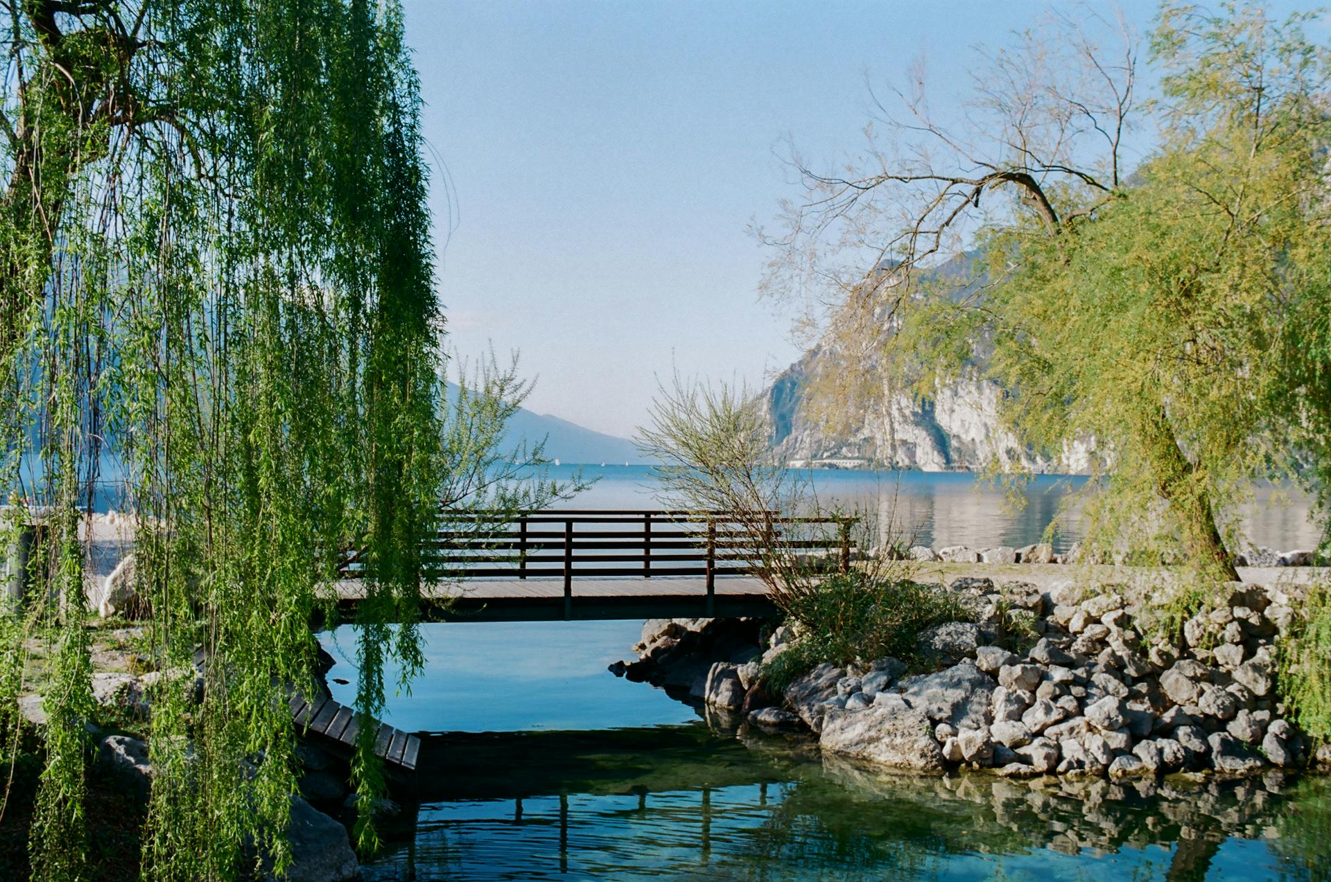 A peaceful scene of a footbridge by Lake Garda surrounded by lush greenery and rocky landscapes.