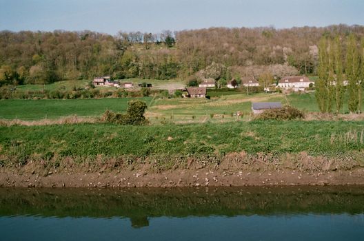 Aged residential cottages located in settlement near green trees on grassy ground near shore of calm lake in rural area of countryside