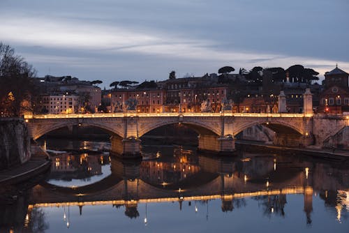 Aged masonry bridge with arches over Tiber River against urban house facades under cloudy sky in evening Rome Italy