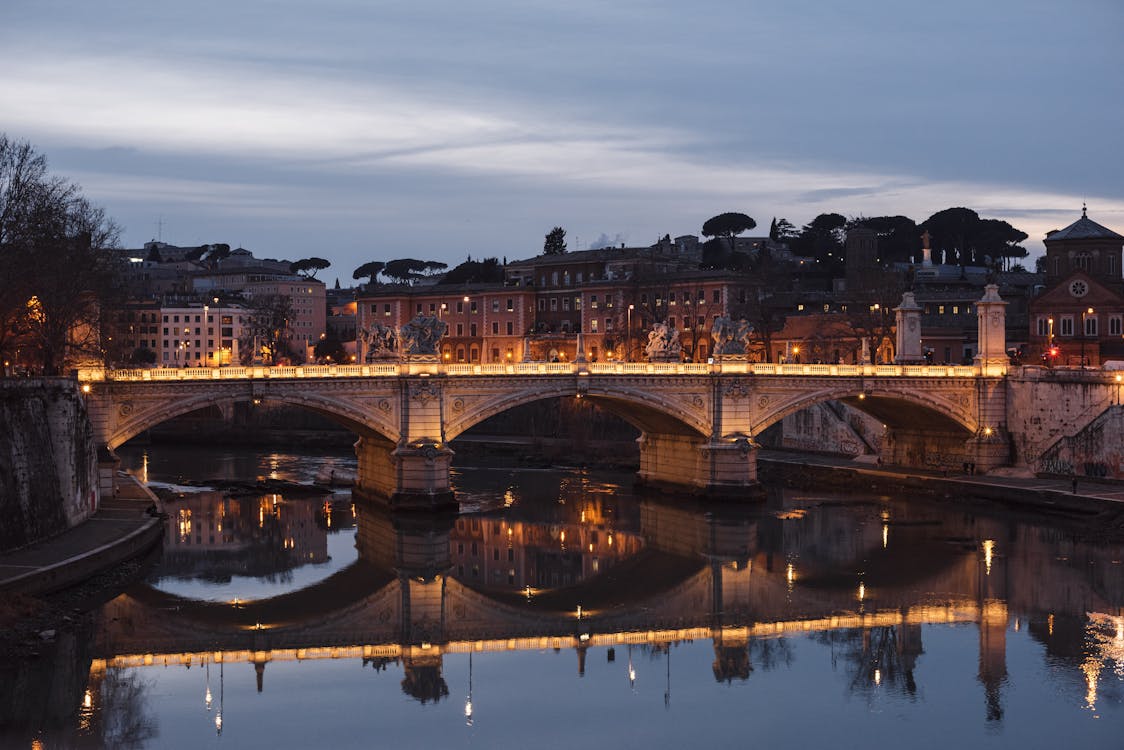 Aged masonry bridge with arches over Tiber River against urban house facades under cloudy sky in evening Rome Italy